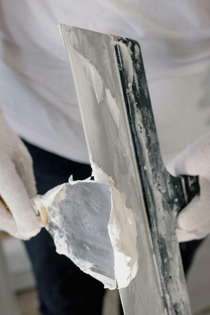 Crop unrecognizable male in gloves holding putty knives while working at new home